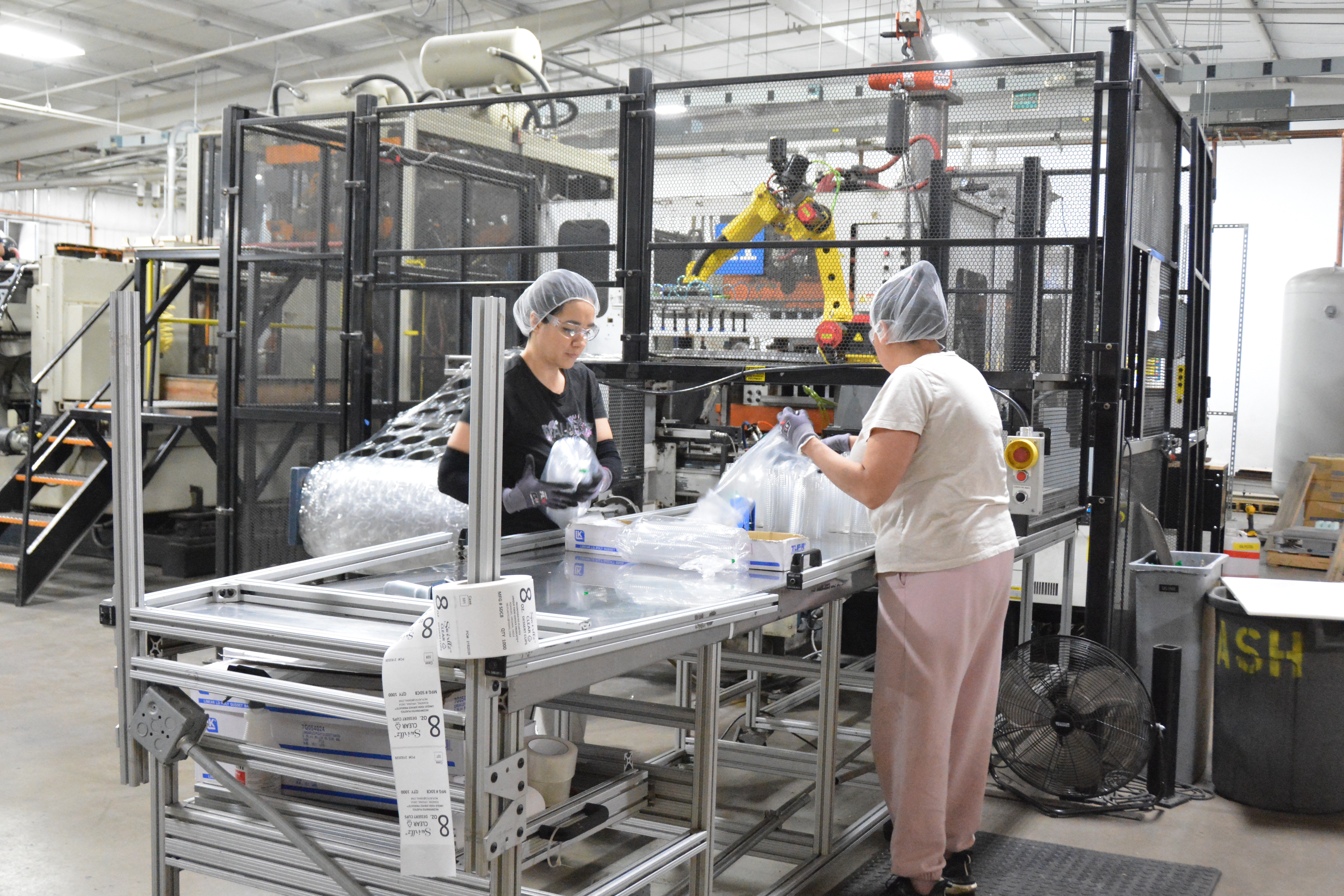 Two MinnTech Plastics workers wearing hairnets and gloves inspecting and packaging clear thermoformed PET clamshells at an end of line quality control station with FANUC robotic cell in background